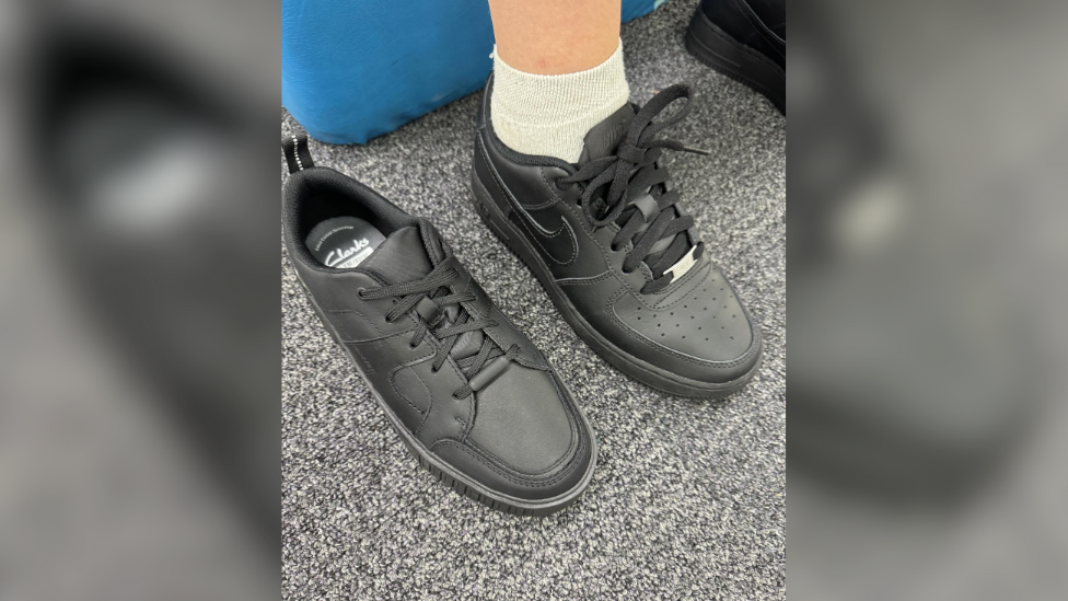 A pair of black Clarks shoes on a grey carpet. The shoes have a black Nike logo on the side and black laces with a metal stud in the centre