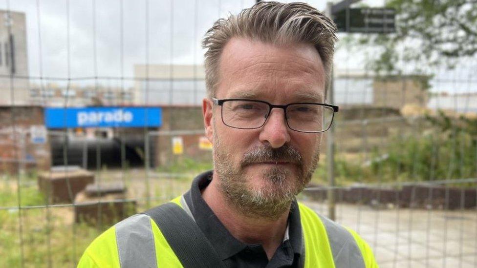 Chris Watts looking into the camera wearing glasses and a fluorescent yellow safety jacket, in front of a building site