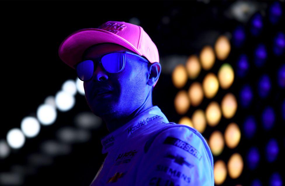 In Pictures: Sporting photos of the week 2 Kyle Larson waits backstage during pre-race ceremonies for the NASCAR Cup Series in Concord, North Carolina. He wears a bright pink cap and a light-coloured racing suit with sponsor logos, under vivid blue lighting and a backdrop of glowing circular lights.