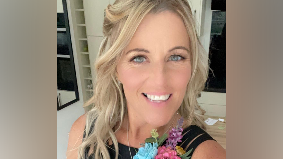 A woman with blonde hair smiles at the camera. She is wearing a black top with a corsage pinned to it. The corsage has blue, pink and purple flowers in it.