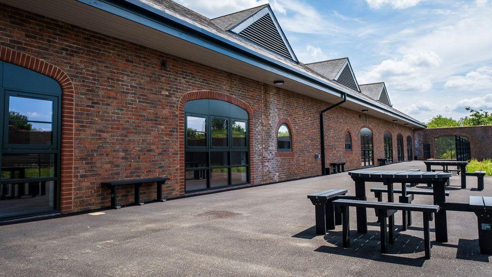 A low-lying single-storey brick built building. It has six large black patio doors and a grey-tiled roof beneath a light blue summer's sky with white clouds. In front of the building is a concrete yard, which has black painted tables and benches.