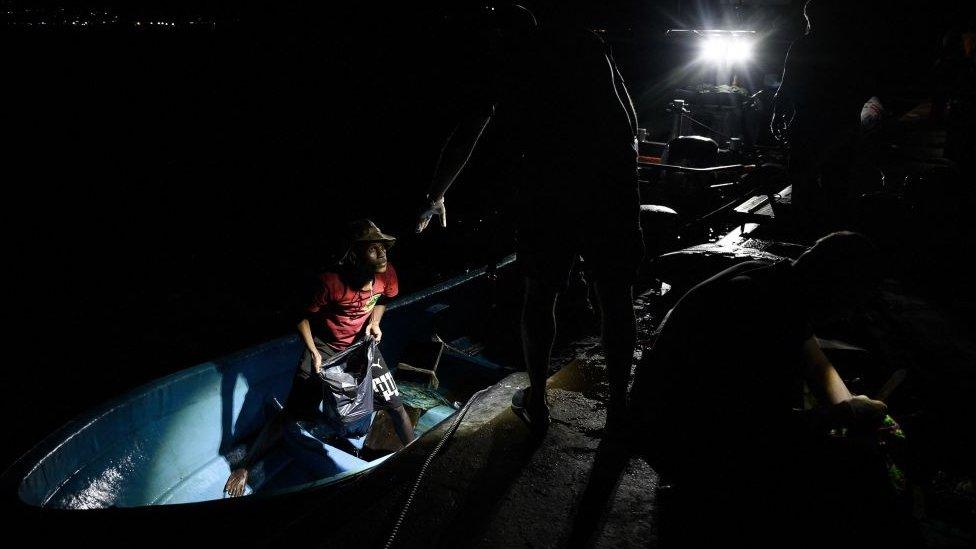 A migrant stands on a boat as he listens to an officer standing on a pier at nighttime