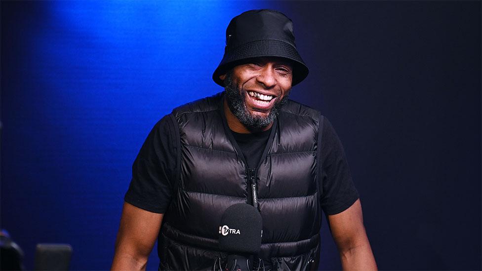 Flowdan, a black man, smiling at the camera. He's wearing a black bucket hat, tshirt and puffer jacket. The background is moody blue lighting.