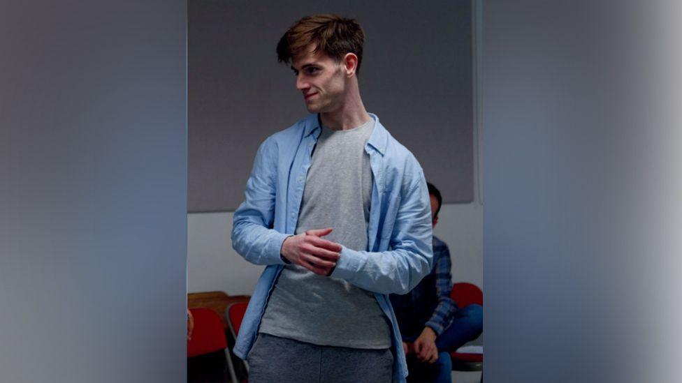 Jude stands and looks to one side during a performance or rehearsal. He is smiling and looks like he is in dialogue with someone else. There are office chairs behind him and someone is sitting on one. He wears a grey t-shirt and open blue shirt, and blue slacks. He has thick short brown hair.