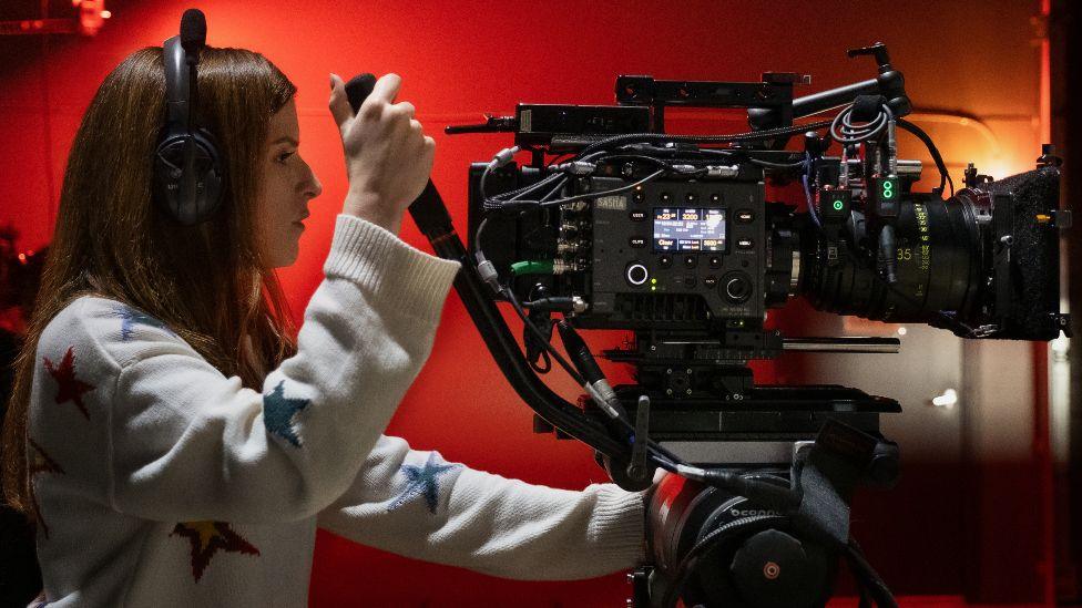 Anna Kendrick on the set of Woman of the Hour. She is wearing headphones and operating a large film camera. 