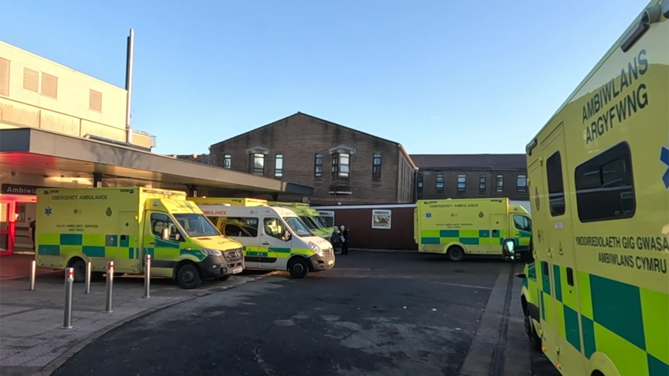 Ambulances waiting outside A&E unit in Swansea. Six bright yellow and green vehicles are in the forefront, with brown buildings behind them