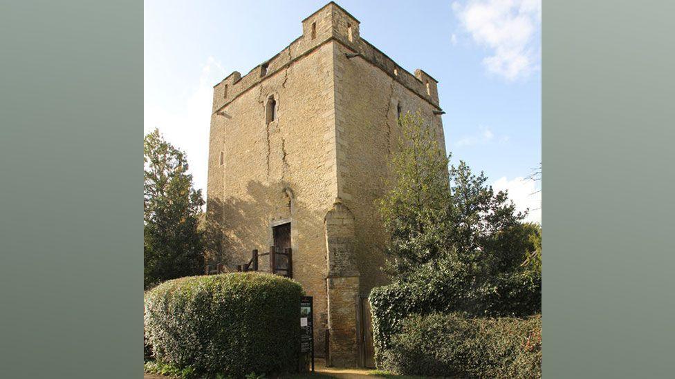 The exterior of Longthorpe Tower, a stone built square-sided tower, which is castellated on the top. An entry to the tower has been cut in a hedge and on the left side of the tower can be seen the top of wooden stairs to its entry which is about a third of the way up the wall. The sky above it is light blue.