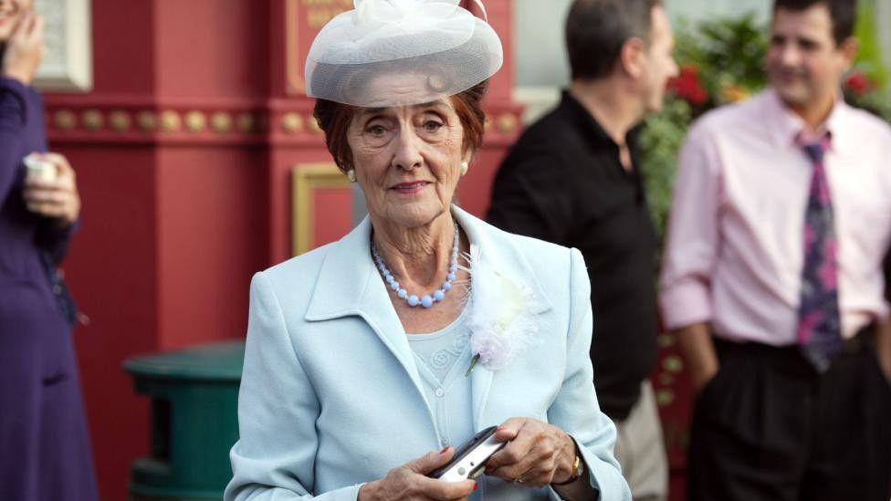 A photo of June Brown playing the character Dot Cotton in Eastenders. She is wearing a pale blue suit and hat and is holding a camera. 