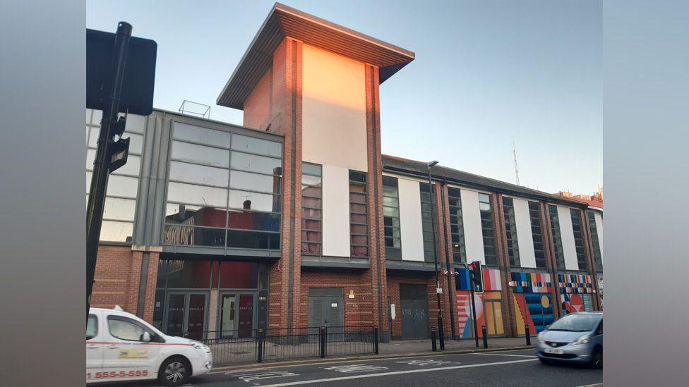 The Sunniside social building is two storeys high. The exterior is a mix of red brick and black glass panelling. There is a colourful mural painted on the left side of the building. 