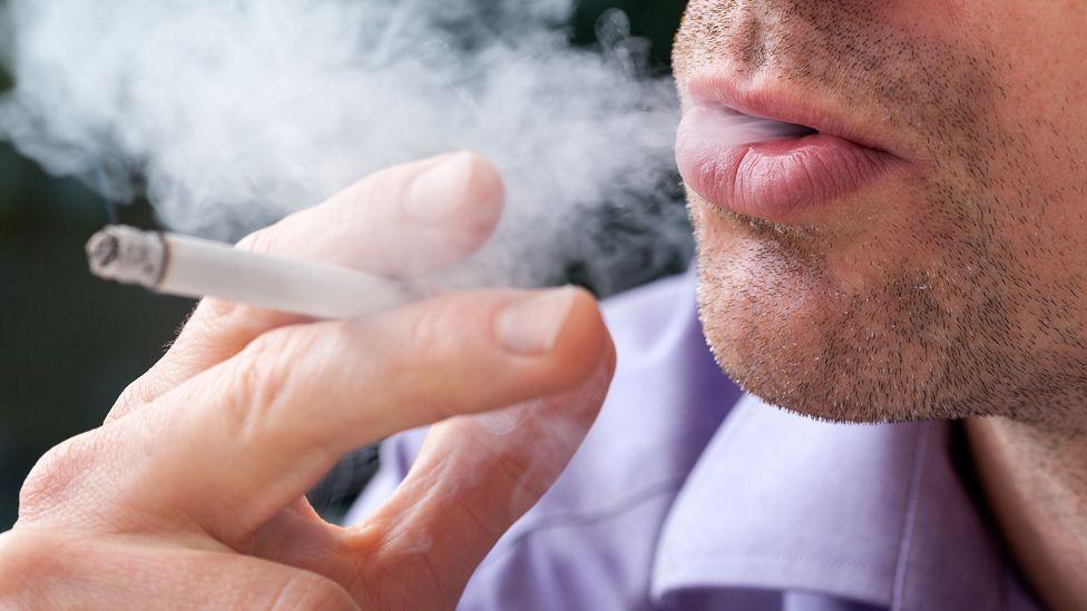 Close-up of a man smoking a cigarette. He has a cigarette in his hand and is blowing smoke out of his mouth.