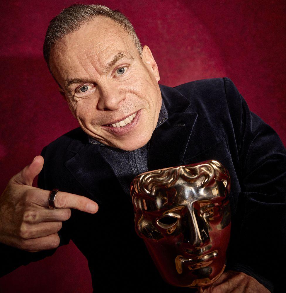 Warwick Davis pointing to his Bafta trophy