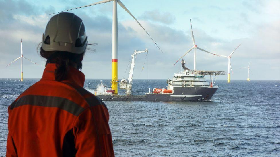 A person wearing orange overalls and a hard hat looks out over the North Sea. There is a ship next to a large wind pylon in the centre of the shot, with other pylons in the background.