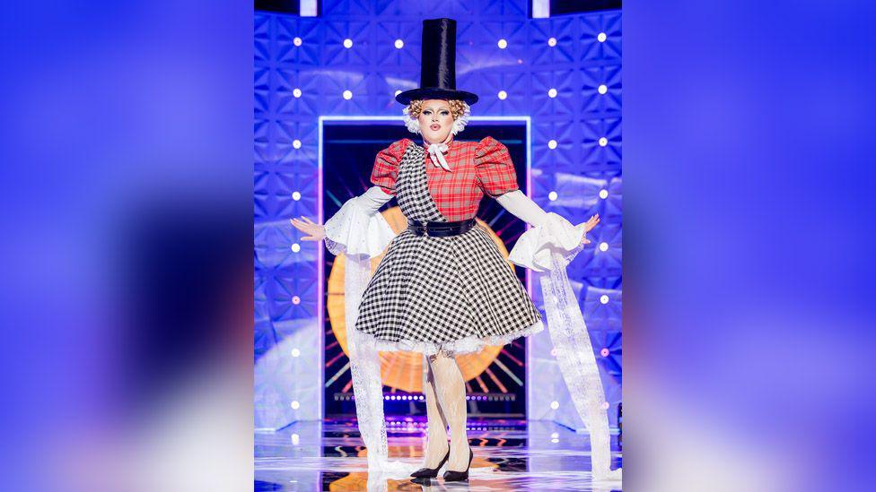 Catrin Feelings is wearing a traditional Welsh hat on top of golden curls, held on with a white ribbon. She is wearing a red tartan puffed-sleeve top, with a black checked sash that goes into a full skirt. She has white sleeves that flow to the floor. 