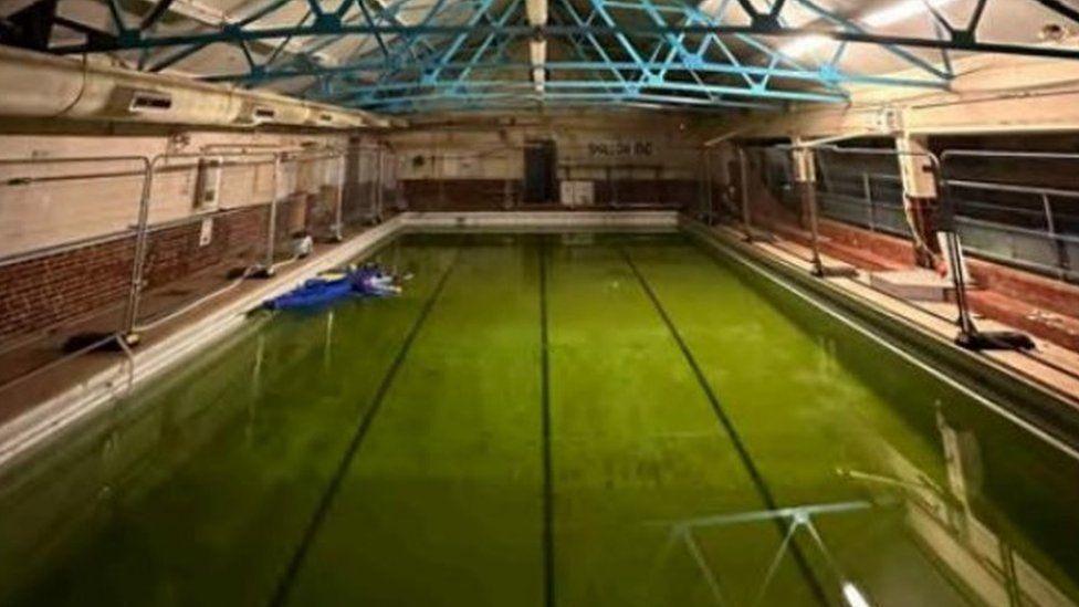 Indoor rectangular pool with four lanes, the water is dark green and metal fencing has been placed around its edge.