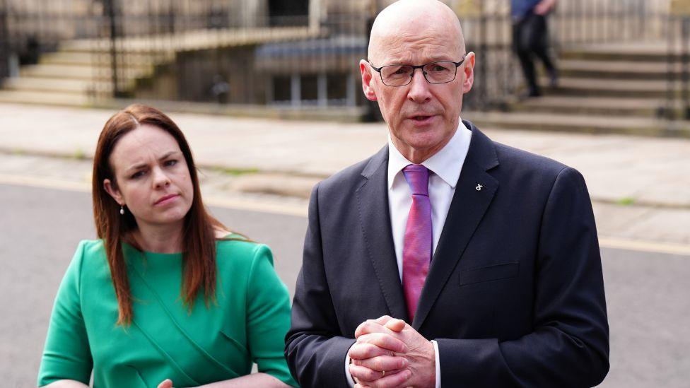 Forbes, with brown hair, stands next to Swinney, a bald man with glasses, on a street in Edinburgh. Forbes is wearing a green dress and Swinney is wearing a dark suit, white shirt and purple tie.