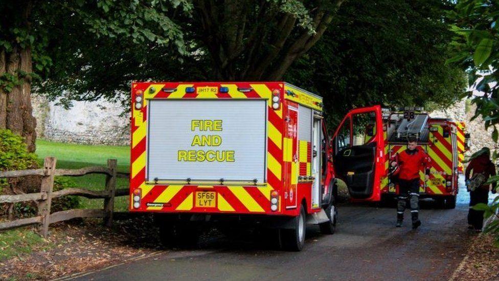 Two fire appliances with castle walls and trees seen behind and firefighter walking towards rear vehicle