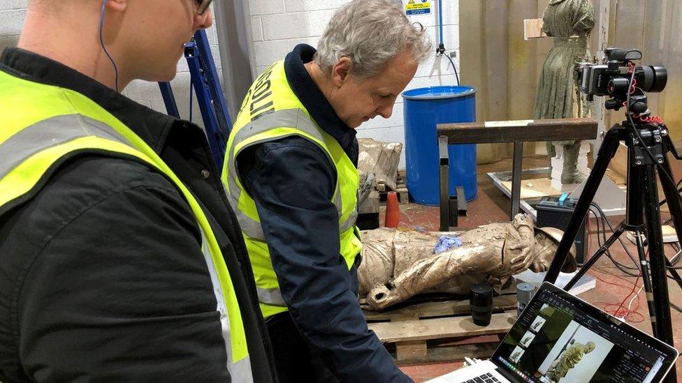 Two men looking at a laptop whilst the bronze cast of a WW1 soldier is laying behind them