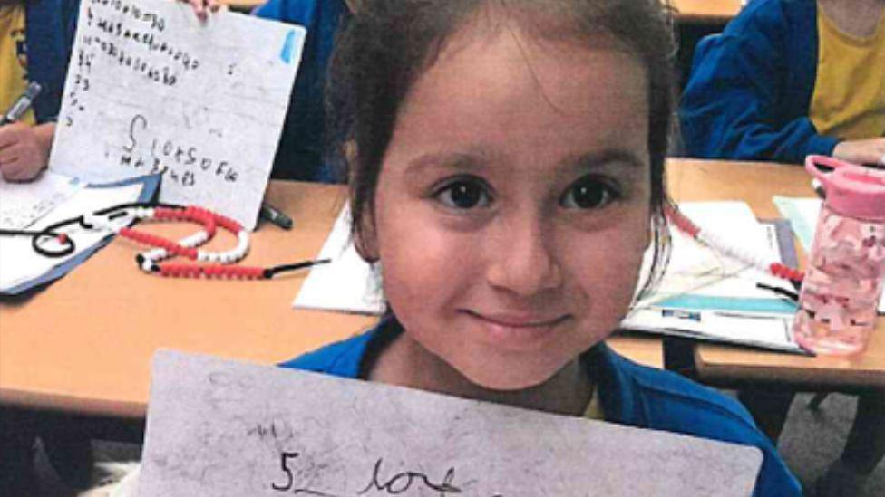 Sara smiling and looking directly at the camera. She is in a school classroom and holding a whiteboard. 