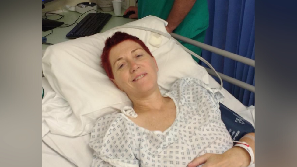 Ms Carey in a hospital bed wearing a clinical white-and-blue hospital gown. She has short red her and is smiling slightly at the camera. Her head is resting on a pillow and she has a paper wristband on her left arm which rests across her stomach. On her upper left arm is what appears to be a blue blood pressure machine. 
In the top right of the image, almost out of shot, is a medical professional at a computer wearing teal-coloured scrubs.