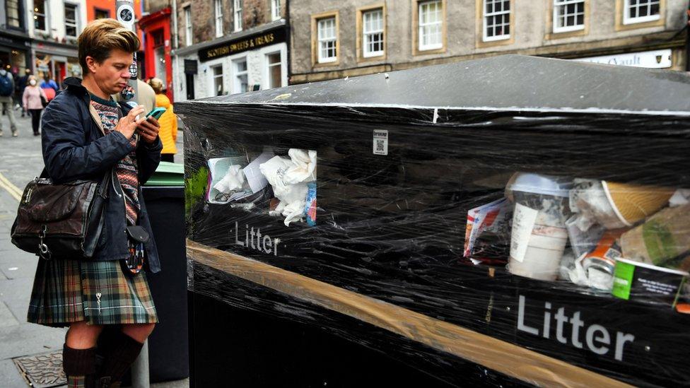 a person stands near full bins in edinburgh which have been wrapped in cling film to stop people throwing their litter away