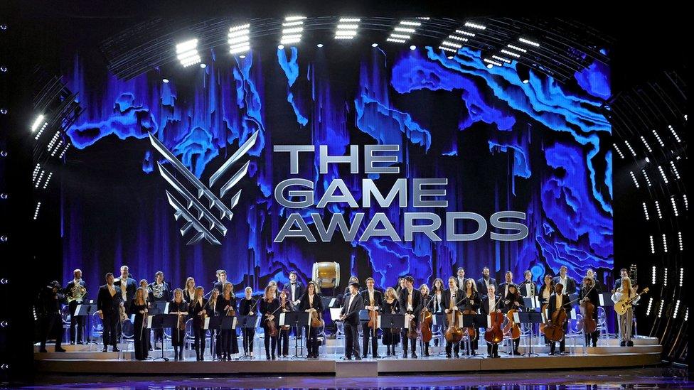 About 40 members of an orchestra holding a variety of instruments line up on stage at the Game Awards. The backdrop is the event logo - an abstract set of diagonal lines forming a V shape - and The Game Awards in large, silver capital letters. The backdrop is a blue-hued, swirling pattern that resembles the rocky interior of a cavern. The stage is so large that the orchestra appears quite small by comparison even though there are three-dozen members.