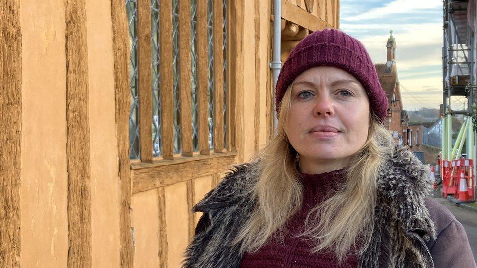 Bernadette Ashcroft wearing a coat and a Beanie hat outside one of the Tudor buildings, typical for the scenery in Lavenham.