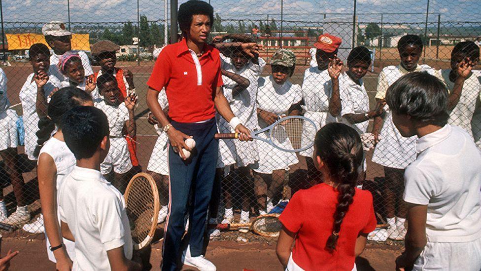 Arthur Ashe in red shirt and navy blue tracksuit bottom and white tennis shoes, holds three white tennis balls in one hand and a grey tennis racquet in the other as he talks to children in front of him during a tennis clinic in Soweto. Others are watching from behind a tennis court fence - November 1973. 