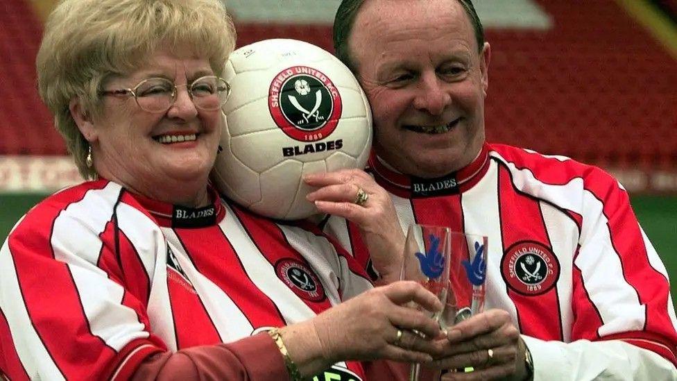 A couple dressed in red and white Sheffield United shirts hold champagne flutes bearing the National Lottery logo aloft. A football bearing the Sheffield United badge is in between their heads.