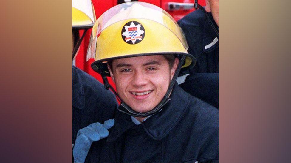 Actor John Alford playing the part of a firefighter: He is wearing  a yellow helmen, with a star-shaped badge, a blue coat and smiling.