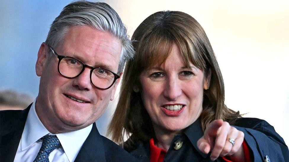 Sir Keir Starmer and Rachel Reeves with their heads together look towards the camera. Reeves is pointing with one hand