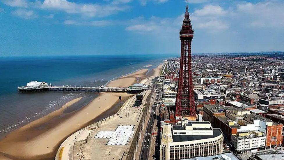 A general aerial view of Blackpool showing the tower and the town to the right, and the beach to the left with Central Pier. The sky is blue with a few white clouds.