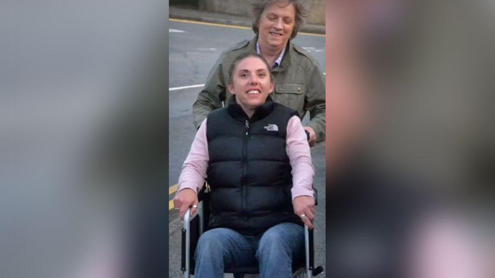 Jules King is sat in a wheelchair. She's wearing a black gilet over a pink sweater and blue jeans. She has brown hair in a ponytail and is smiling. There's a person behind her in an olive green jacket with medium length grey hair smiling