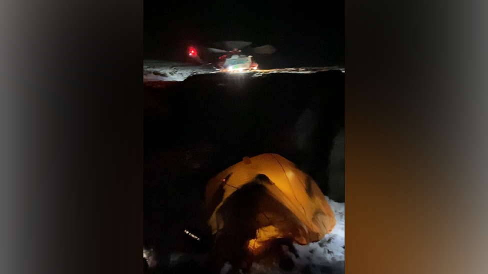A helicopter at night on a snowy mountain with a yellow tent in tghe foreground