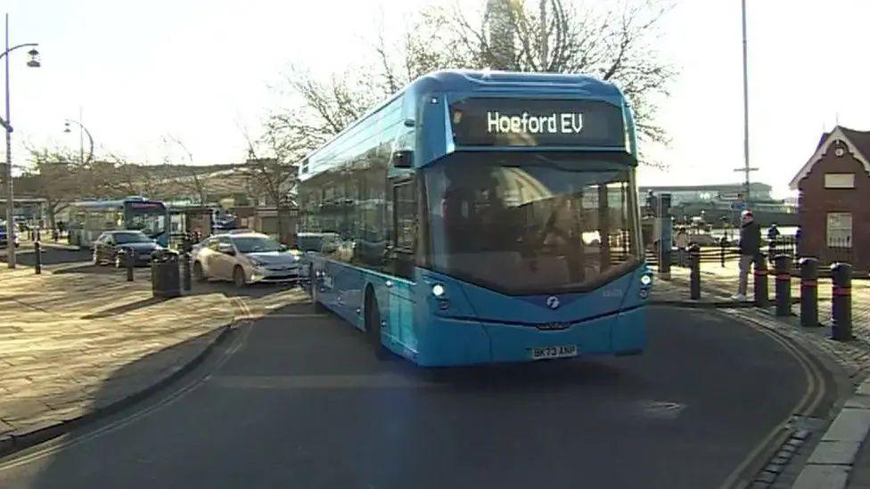 A blue bus turning a corner in the road pulling out of a bus stop, with Hoeford EV written on the front of it.