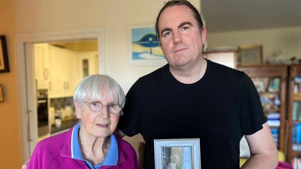 A man wearing a black t-shirt and holding a framed photo of a woman. He has his arm around a woman wearing a purple and blue shirt who is standing to the right of him.