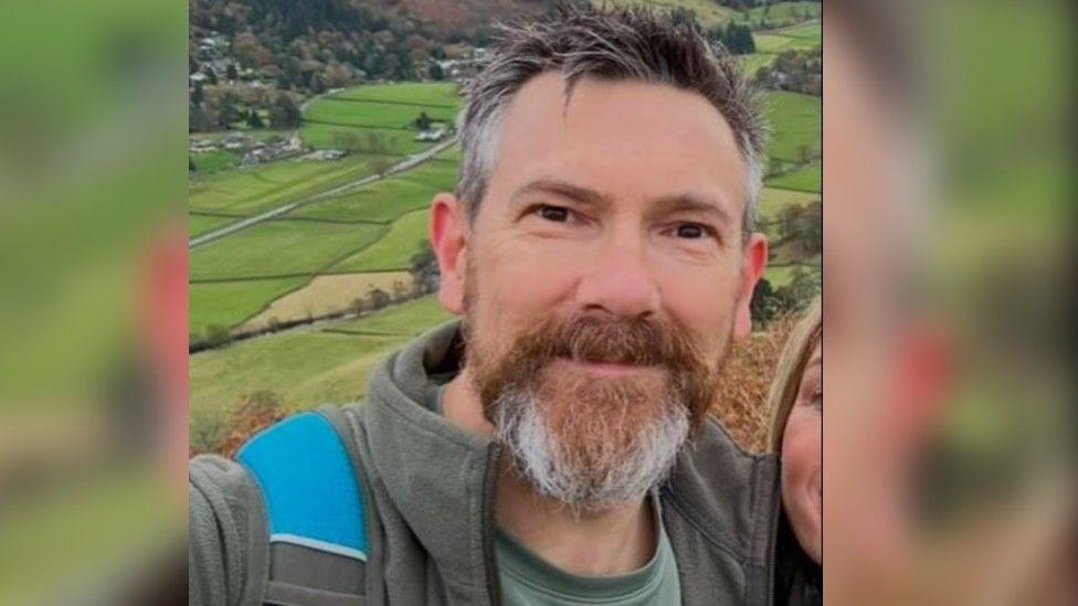 Richard Mason smiling looking at the camera. He is wearing a green t-shirt and a green zip-up fleece. He had a backpack on his shoulders that has a black and blue strap. He has a brown and grey bushy beard and dark hair. 