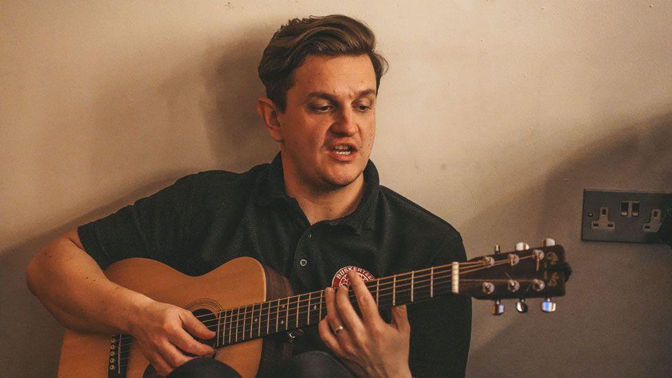 Andrew Small in a black polo shirt strumming a guitar and singing