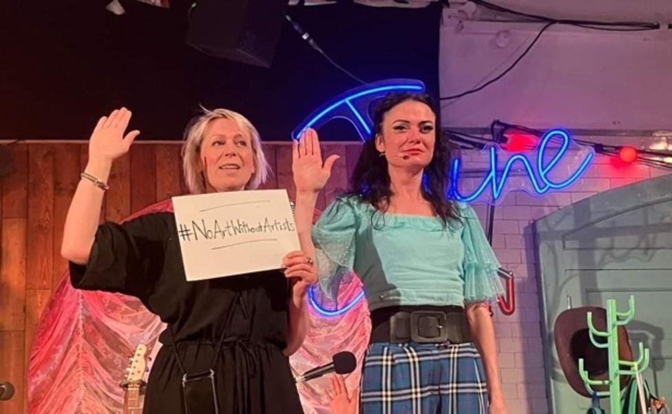 Two women from a theatre production standing side-by-side on stage protesting arts cuts