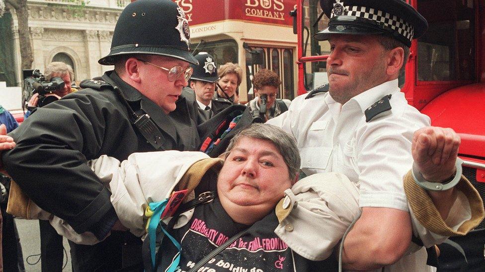 A woman is carried from in front of a bus by two policemen on Whitehall