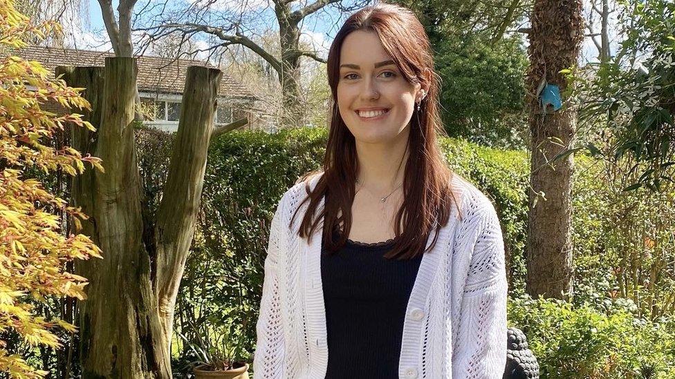 Emily has long brown hair, tied back in a half-up, half down style with bangs. She's wearing a cream cardigan and black vest top. She's standing in a garden. You can see autumnal trees, hedges and the top of a house in the background.