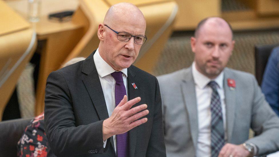 John Swinney, a bald man with glass, wearing a dark suit and purple tie, stands to speak in the Scottish Parliament 