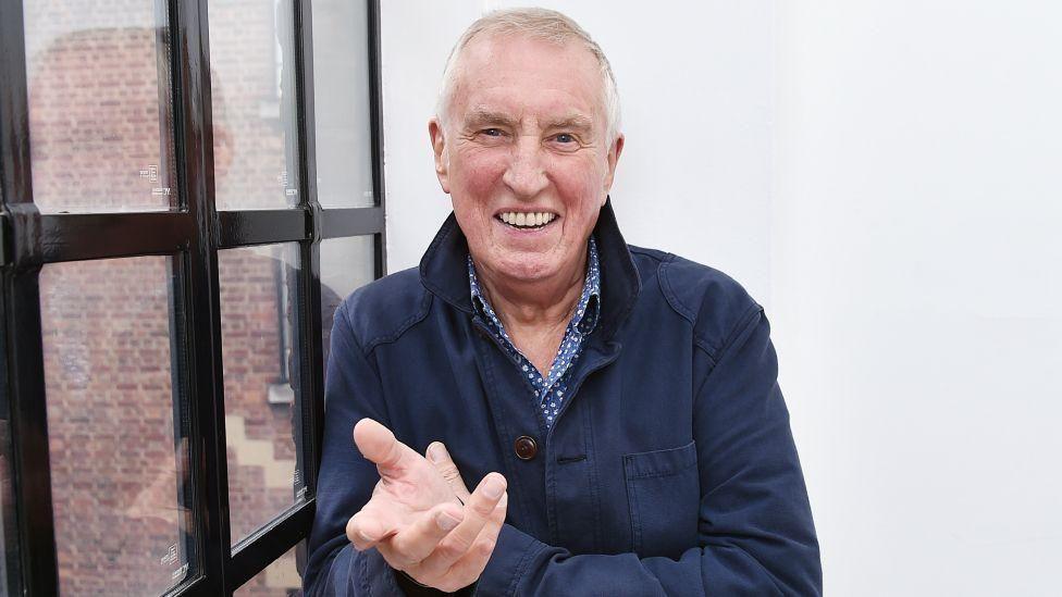 Johnnie Walker, with white hair, a blue jacket and smiling at the camera by a window.