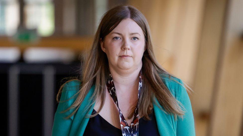 A lady with a serious expression pictured walking down a well-lit corridor in the Scottish Parliament building. She has long brown hair and is wearing a green jacket, black top and a patterned neck scarf.