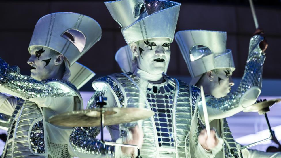 Three performers wearing white face paint and white and silver costumes are stood with their backs to each other. They are playing drums and symbols.