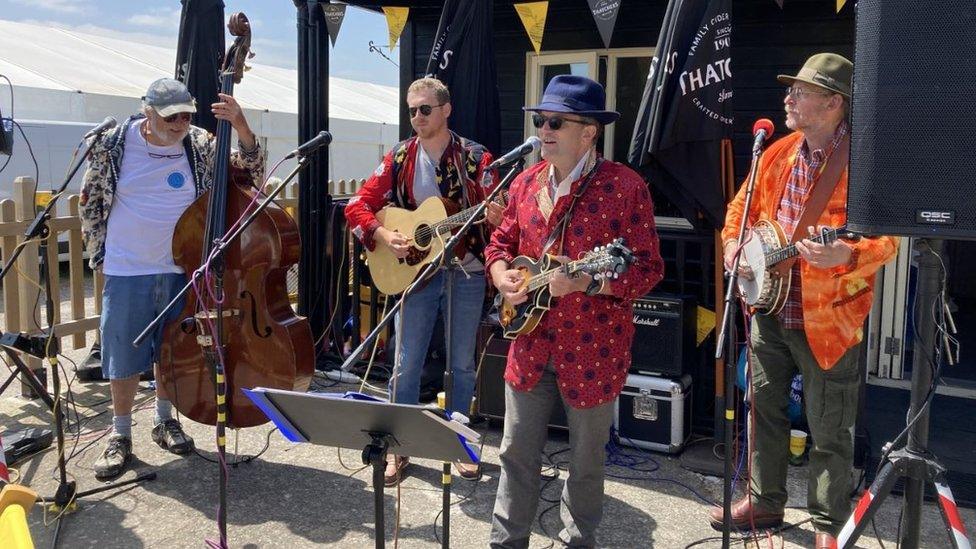 A band of four men sing and play in front of microphones, two have guitars, one has a banjo and one had a double bass