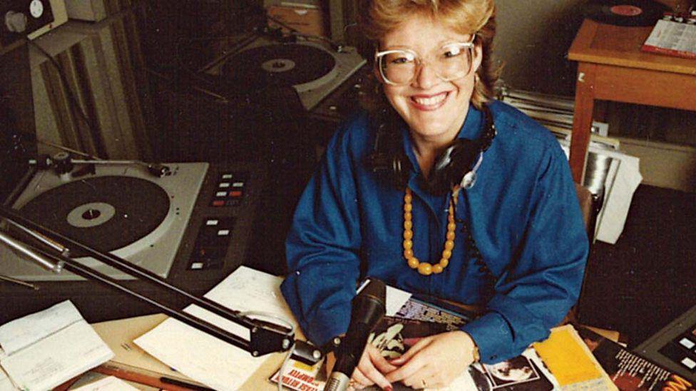 A woman sitting at a radio desk with a microphone and turntable and magazines over the desk. She is wearing a blue shirt and headphones round her neck and smiling up at the camera