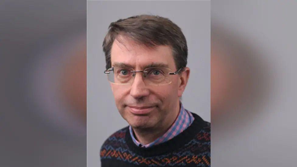 David Rouane headshot in front of a grey background. He has short brown hair, glasses and is wearing a colourful knitted jumper.