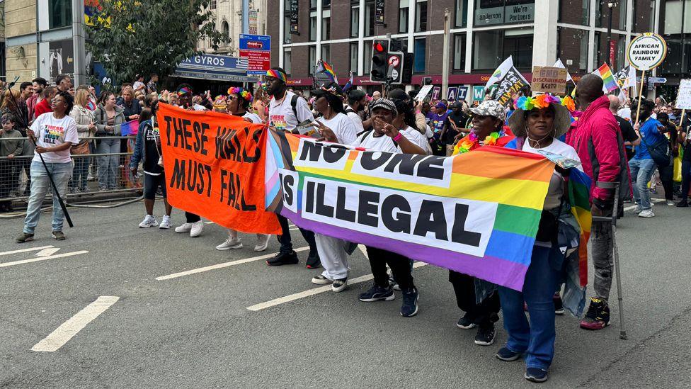 A group wearing rainbow-garlanded hats and rainbow headbands, carry a banner reading "No one is illegal" and "These walls must fall" in the Manchester Pride parade