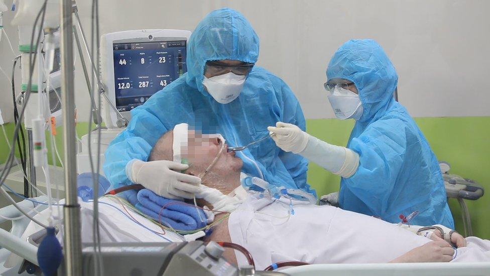 Stephen Cameron, face blurred, is fed by masked staff in a hospital bed