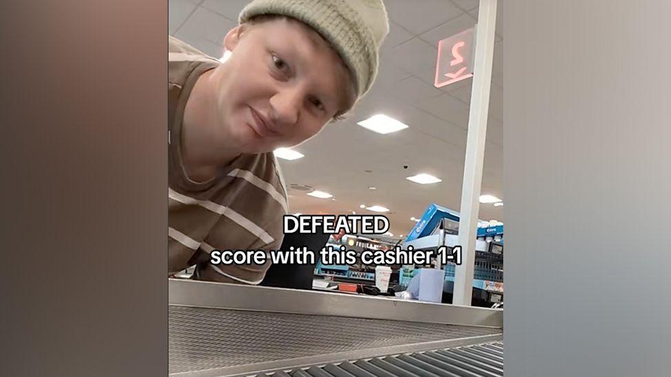 Leroy Lupton at the Aldi checkout wearing a woolly hat with the text on the image "defeated score with this cashier 1-1"